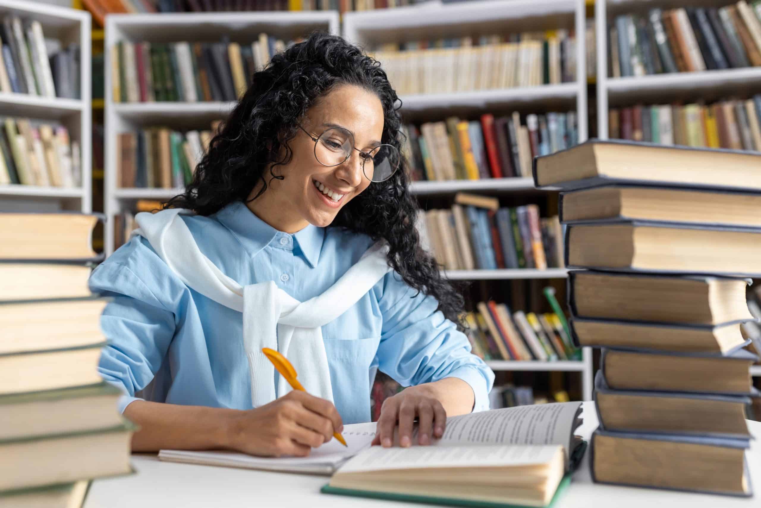 jovem mulher sorrindo sentada em uma biblioteca, rodeada de livros e escrevendo em um caderno