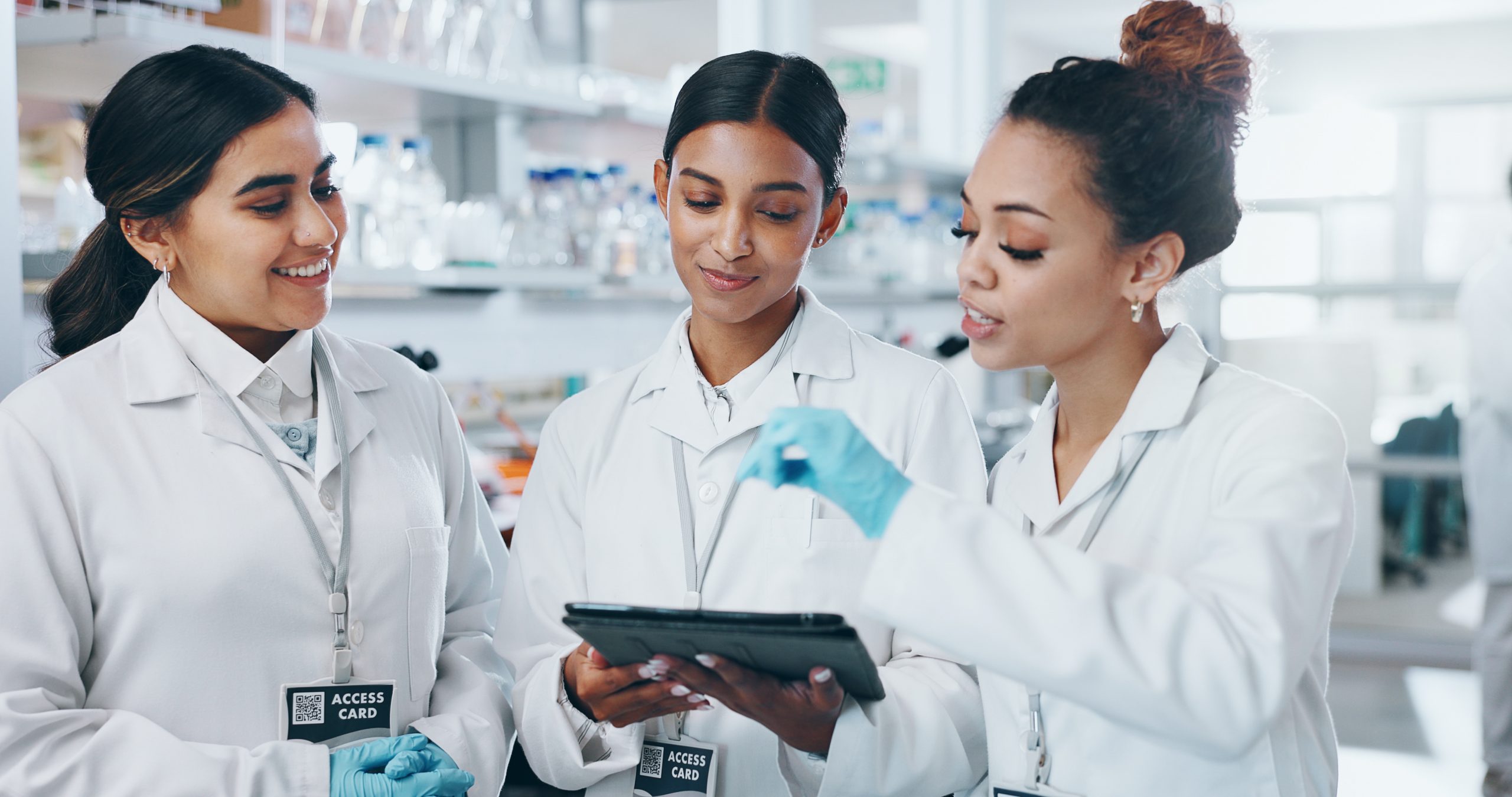 Três jovems meninas em ambiente farmacêutico, vestidas com jalecos e outros equipamentos de uso pessoal devidos para a área
