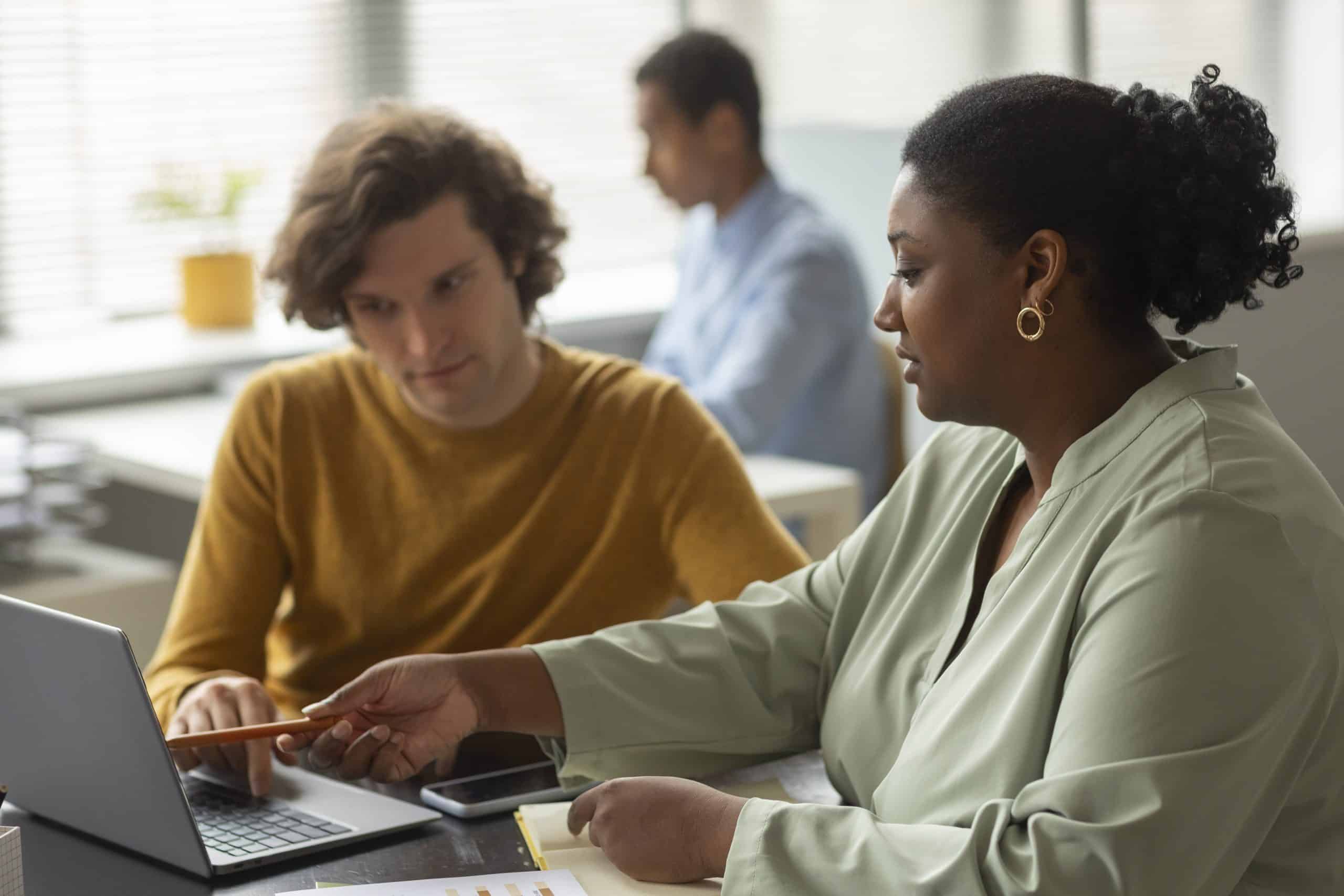 Uma mulher e um homem conversando em frente ao computador. A mulher pode ser considerada uma professora por estar apontando algo como ensinamento no computador