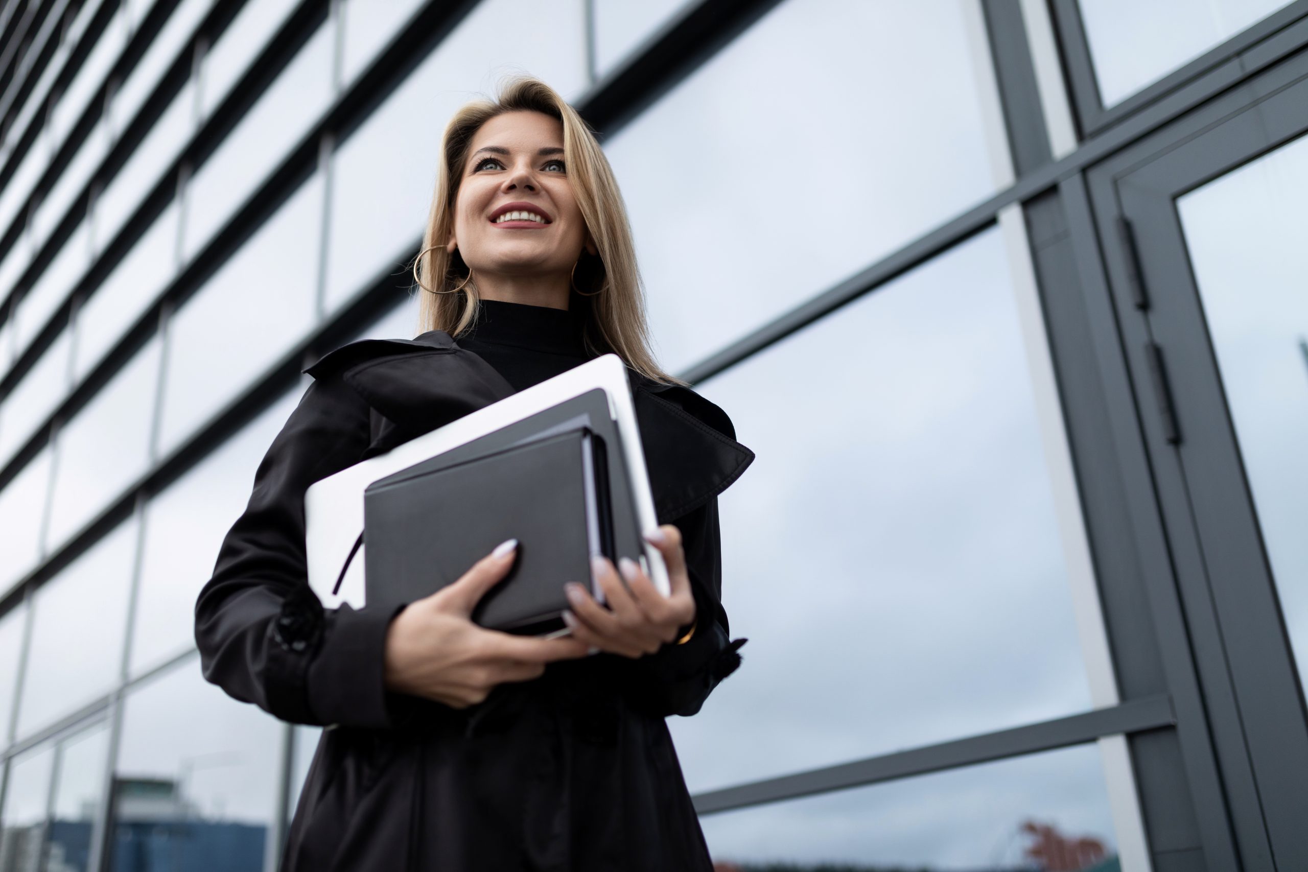 mulher segurando agenda, documentos e notebook em frente a um edifício espelhado