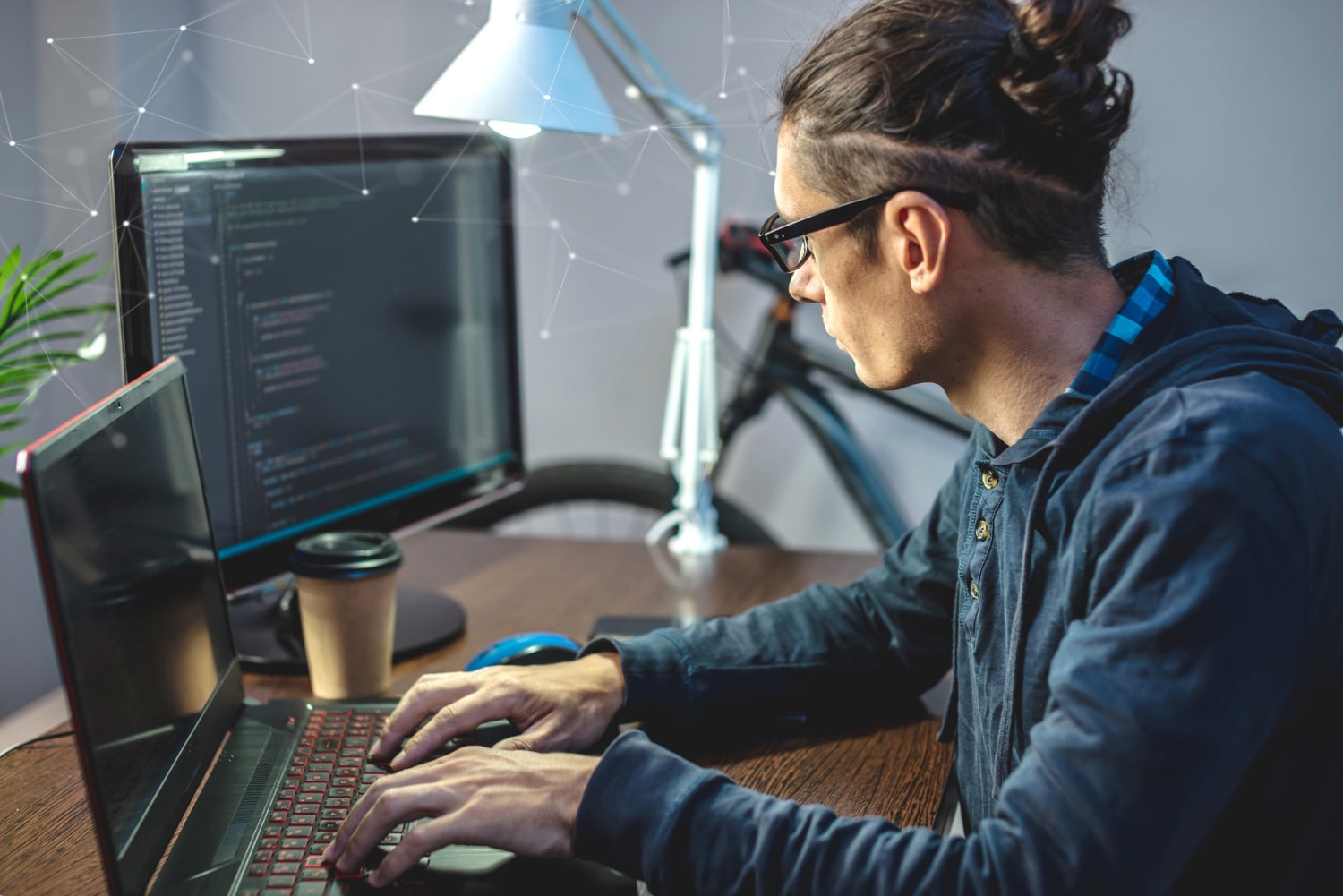 Jovem adulto sentado em frente a um notebook e usando um monitor auxiliar para programar.