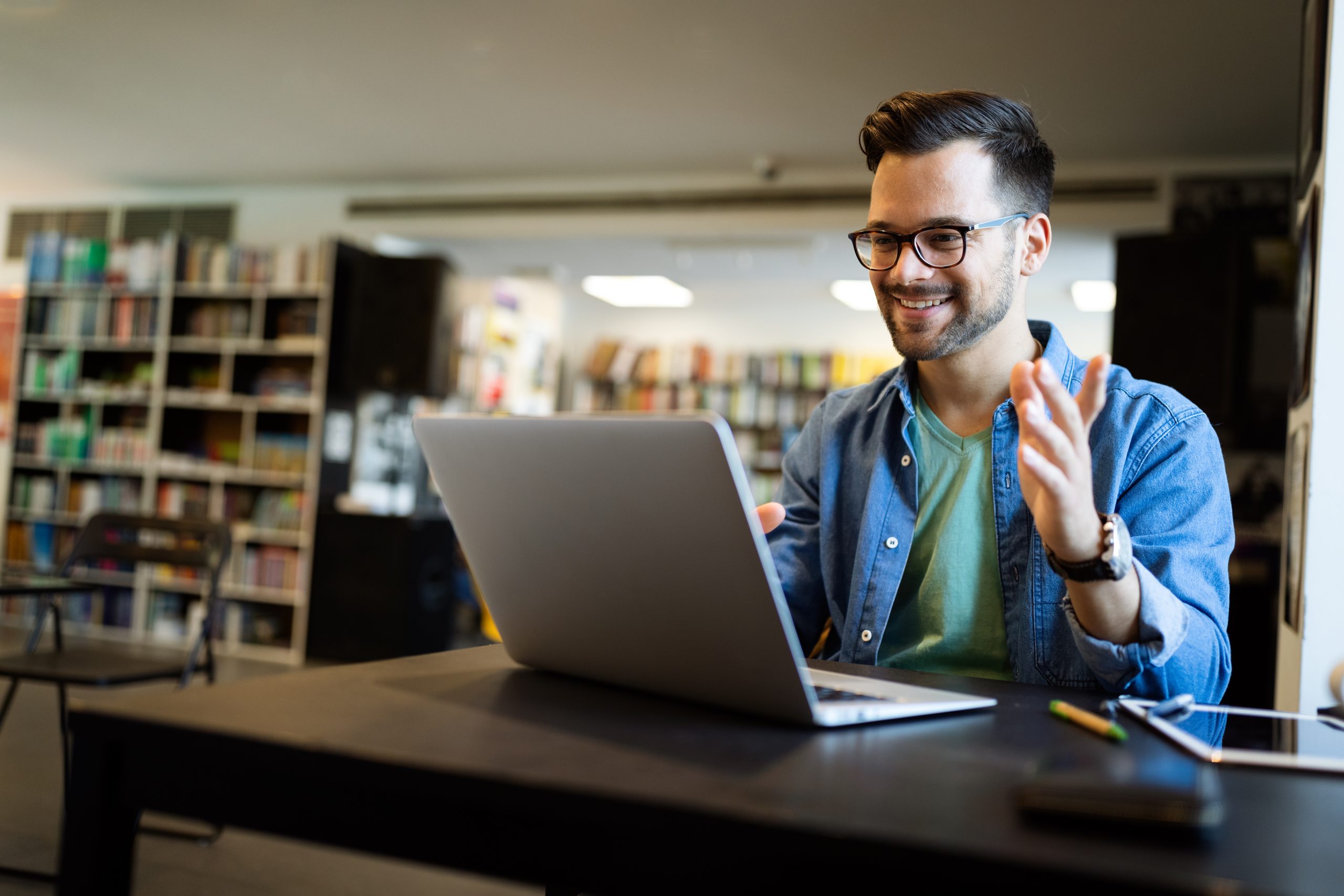 Jovem adulto sentado em uma biblioteca participando de aula online pelo notebook.