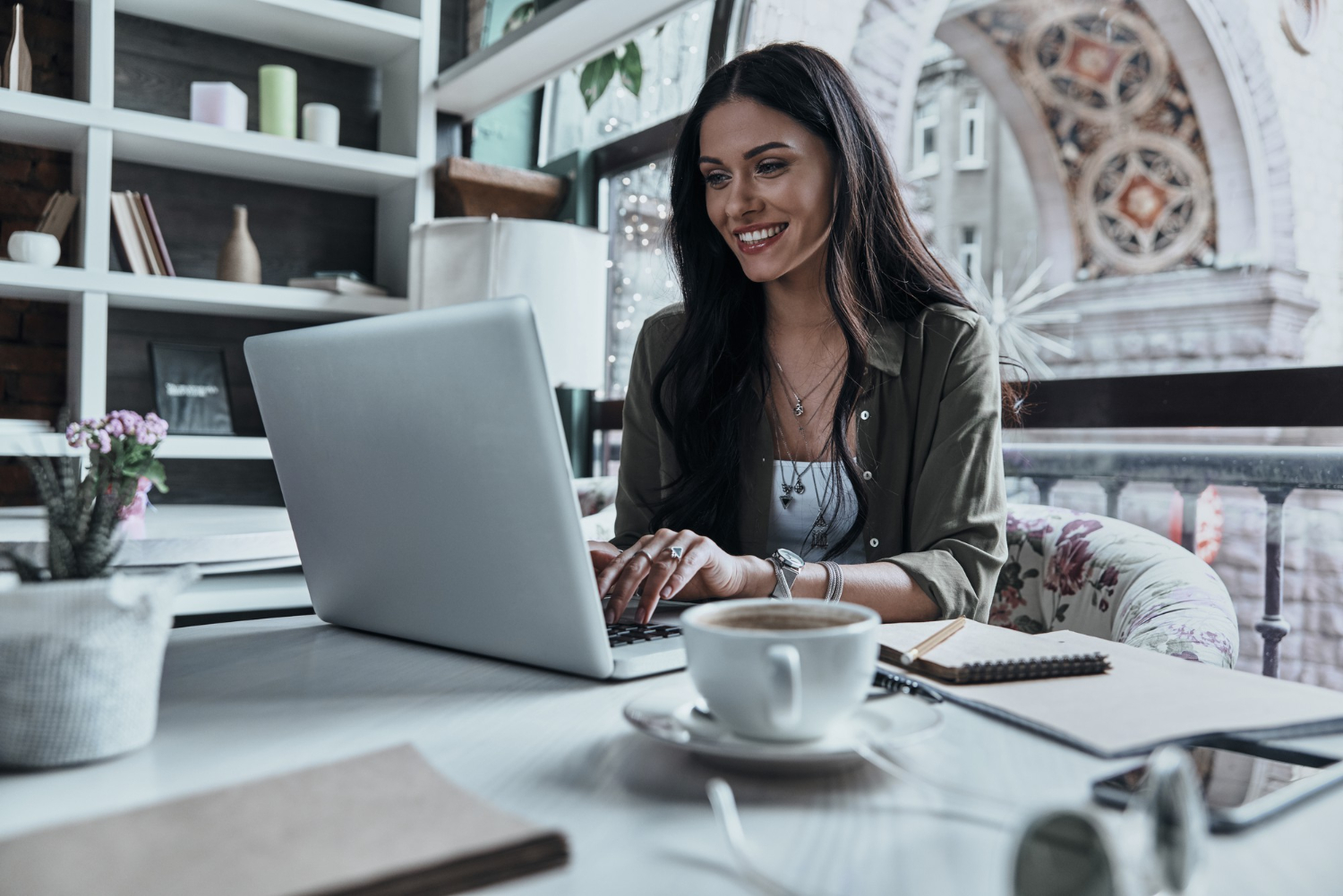 Mulher em um escritório, sentada em seu ambiente de trabalho. Atrás dela há uma janela. Ela está sentada sorrindo para notebook. Ao seu lado uma xícara, cadernos e materiais de escritório sobre a mesa que está usando.