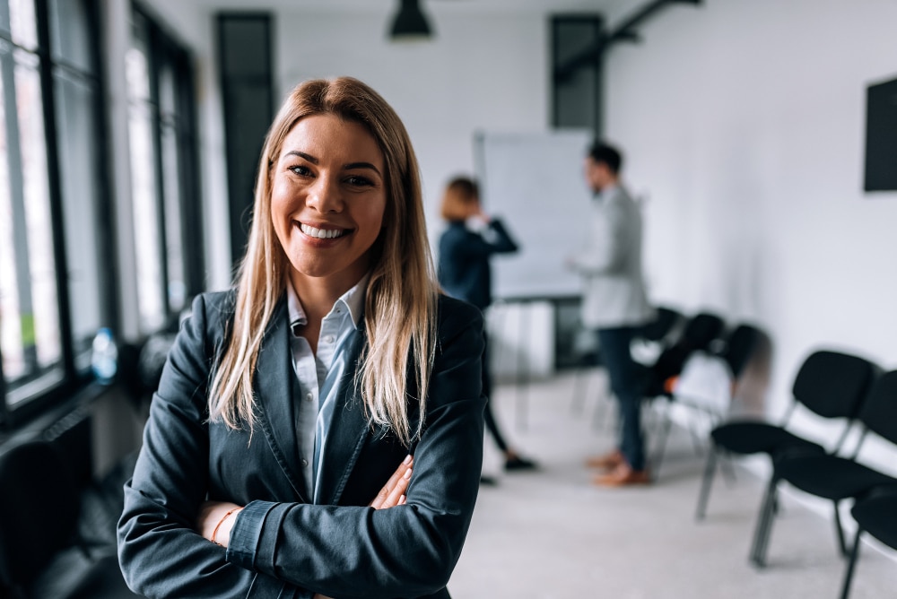 mulher em ambiente corporativo. Ela está sorrindo para a câmera com os braços cruzados