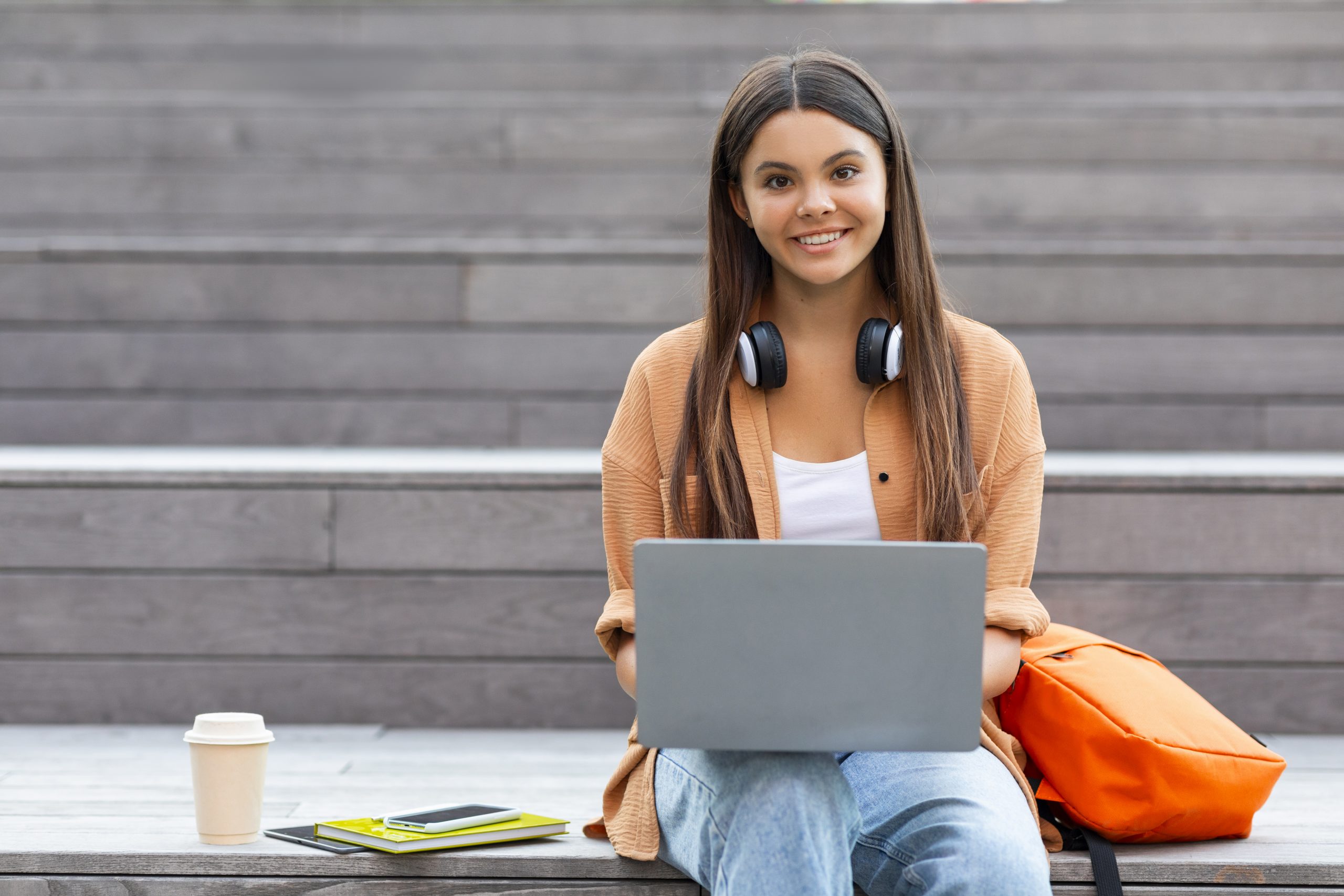Jovem mulher sentada na escadaria da faculdade com seu notebook aberto no colo, ao seu lado seus materiais da faculdade, uma mochila e um copo de café.