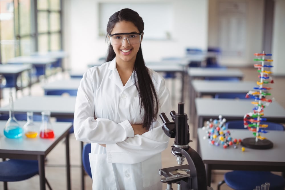 jovem adulta de jaleco branco em sala de aula com equipamentos de pesquisa. Ela está de braços cruzados sorrindo para a câmera.