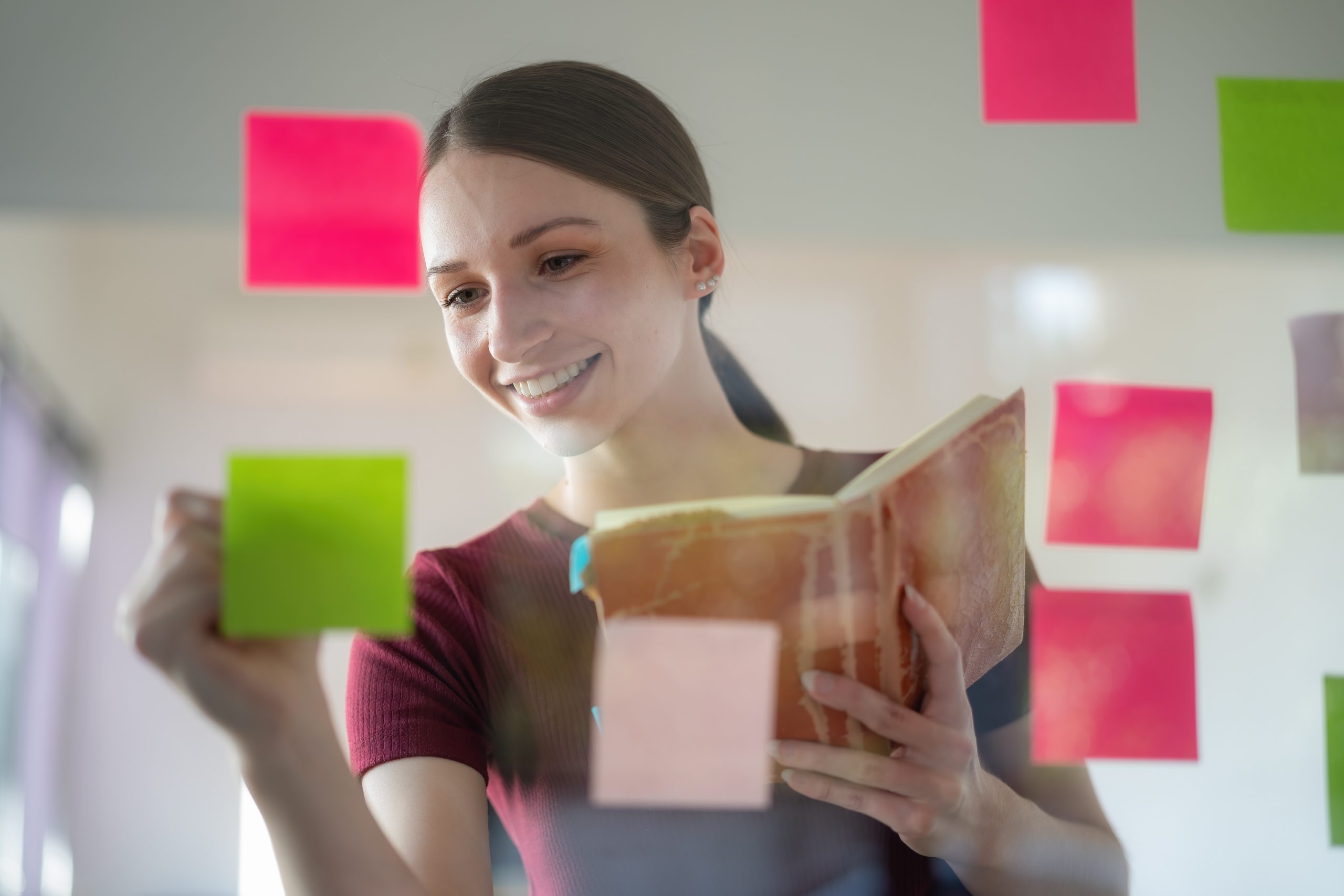 Jovem adulta segurando um livro aberto em mãos. Ela está sorrindo para postit a sua frente que estão colados em um vidro, ao mesmo tempo que faz alguma anotação.
