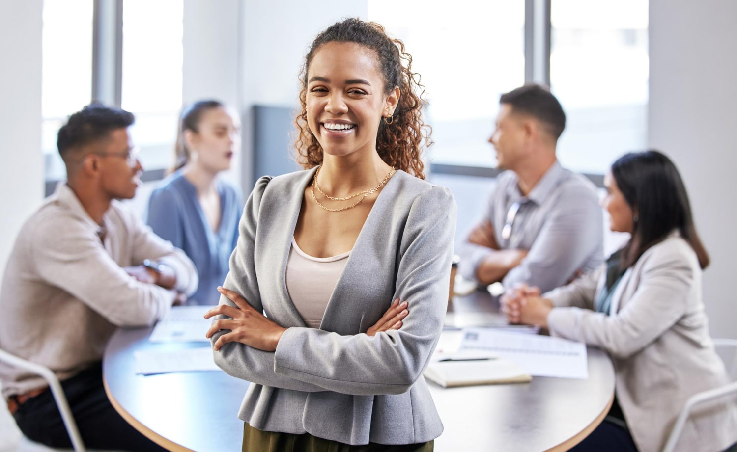 Mulher em pé sorrindo com braços cruzados, ela está em ambiente de trabalho, ao fundo uma mesa de reuniões com outras pessoas conversando.