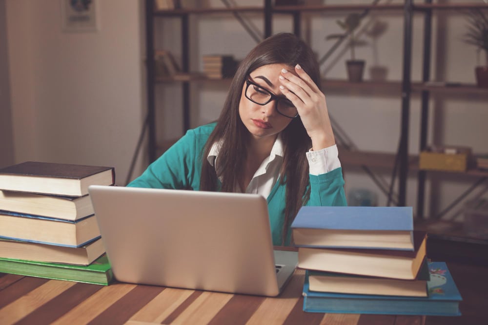 jovem adulta sentada em mesa de estudos, rodeada de livros e com notebook aberto a sua frente. Ela está com expressão de cansaço, com a mão apoiada na mesa e segurando a cabeça
