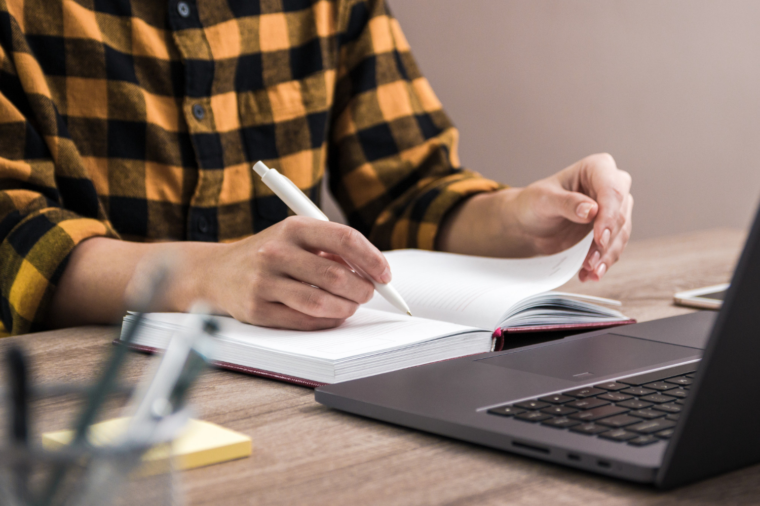 meia imagem de um homem sentado com um caderno aberto a sua frente e um notebook. Ele está fazendo anotações no caderno.