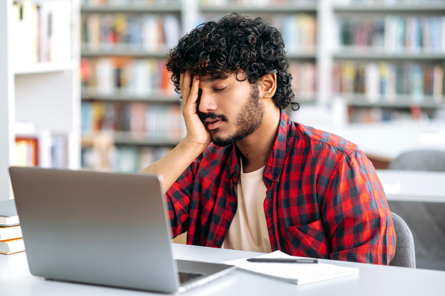 aluno sentado na biblioteca com expressão de cansado em frente ao notebook