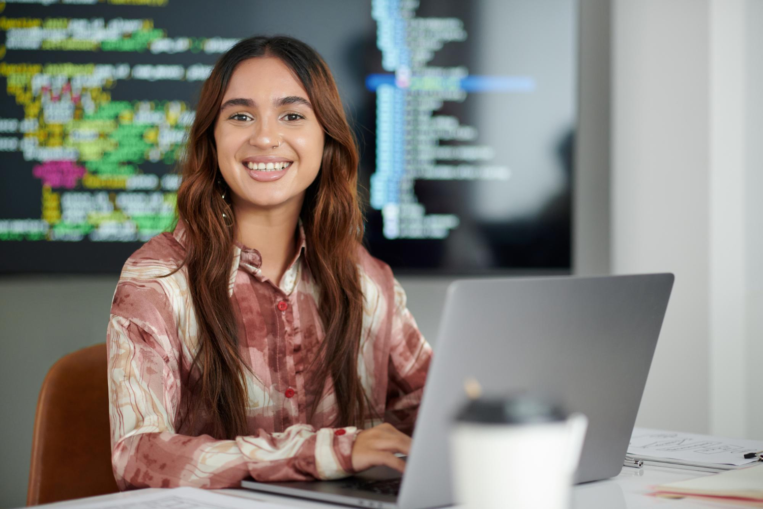 jovem adulta trabalhando com notebook. Atrás dela, uma imagem de códigos de programação