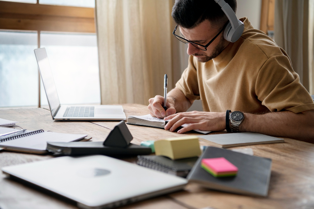 Homem com fone de ouvido em frente a uma mesa, na mesa há notebook e outros itens de escritorio.