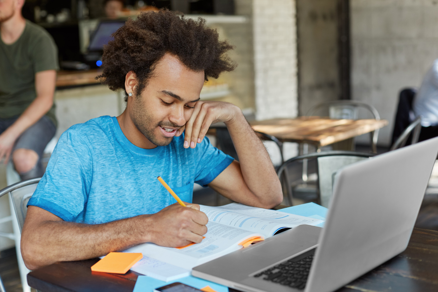 jovem adulto sentado em frente ao notebook com um livro aberto fazendo anotações de estudos