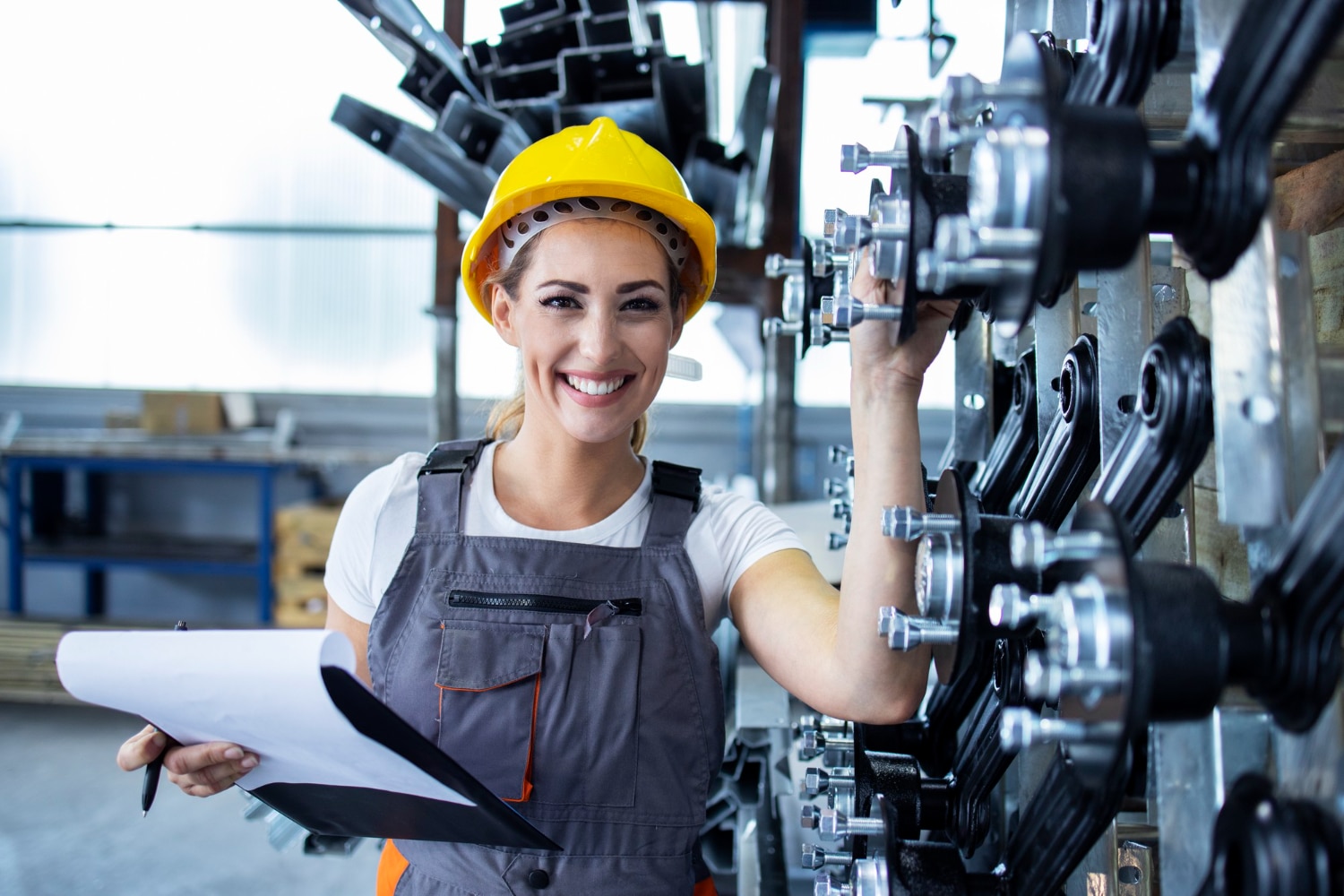 mulher, engenheira mecânica, em fábrica de montagem de equipamentos. Ela está sorrindo para a câmera, usando macacão cinza e capacete amarelo, com uma prancheta em uma das mãos