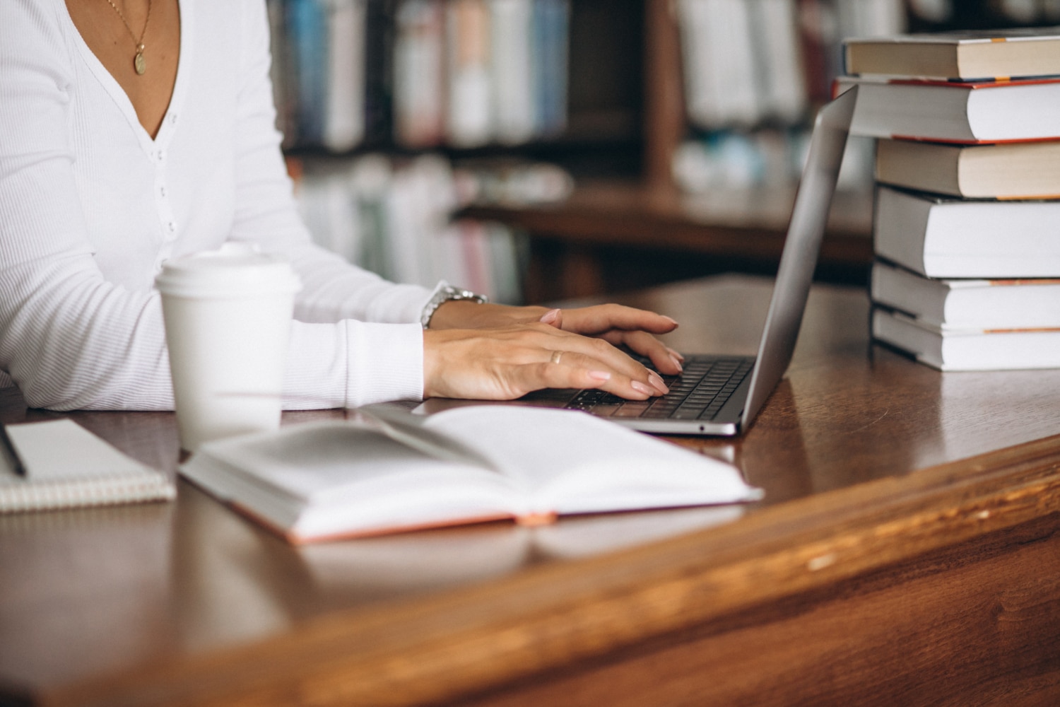 imagem somente das nãos de uma mulher digitando em um notebook sobre uma mesa. Ao seu lado, um copo de café, uma agenda aberta e uma pilha de livros
