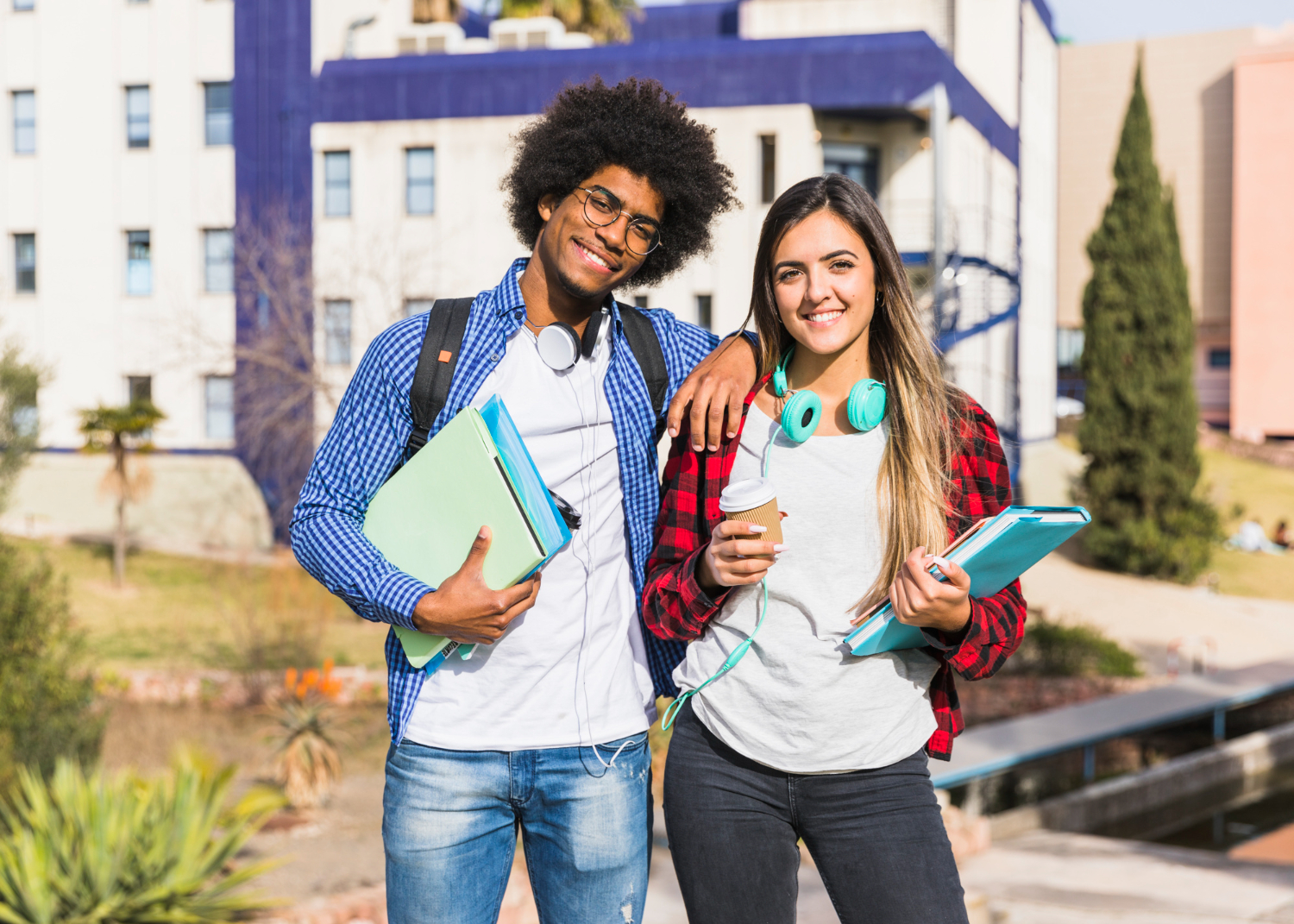 dois jovens adultos lado a lado sorrindo para a câmera. Ele está segurando materiais como pastas e cadernos em uma das mãos e com o outro braço apoiado na menina que segura um copo de café em uma das mãos e um caderno na outra. Eles estão na rua, em frente a um prédio de faculdade.
