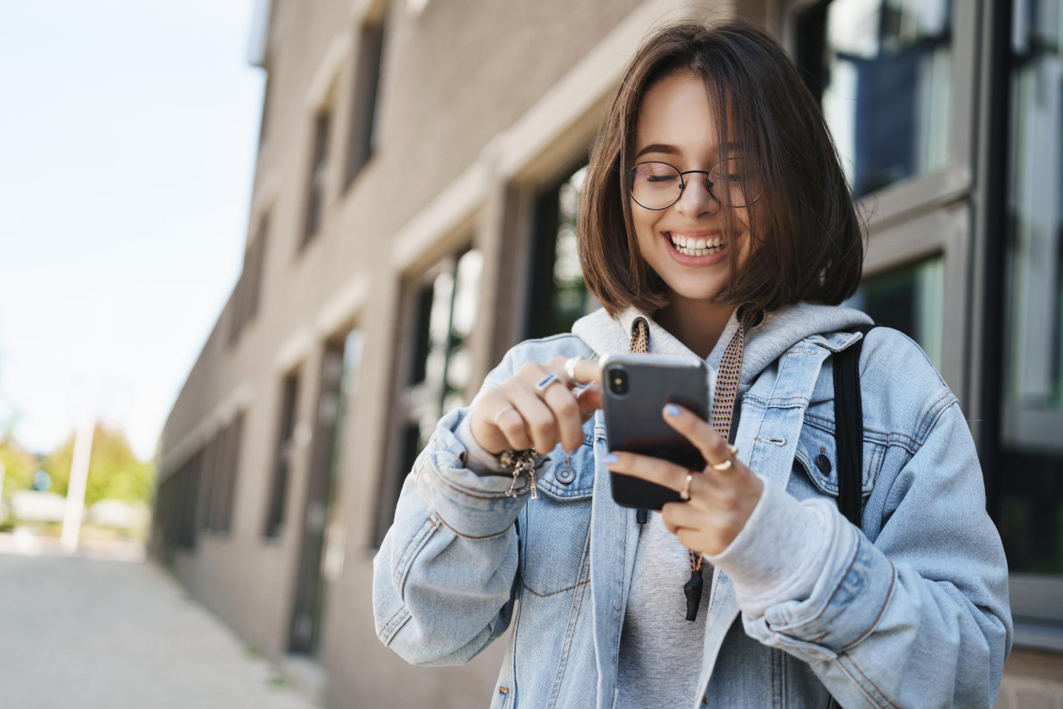 jovem adulta em frente a um prédio que remete a faculdade. Ela está sorrindo ao olhar para o celular. Sua vestimenta é de jovem universiária, com jaqueta jeans e mochila