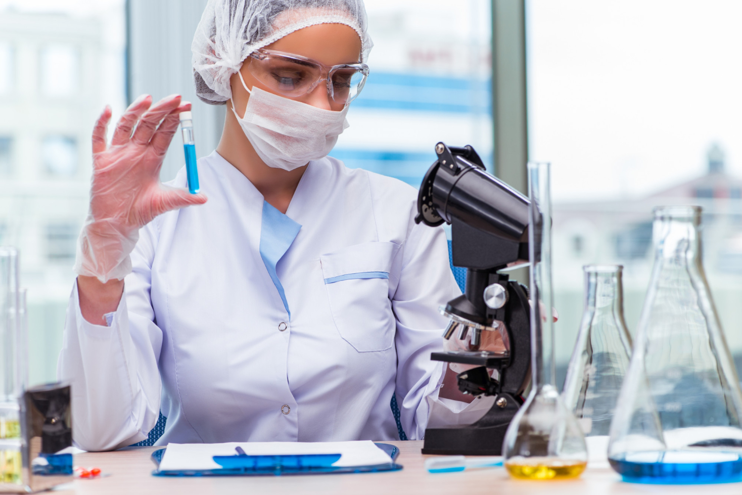 mulher paramentada trabalhando em laboratório. Em uma das mãos um tubo de análise, a sua frente um microscópio, uma prancheta e outros tubos de ensaio