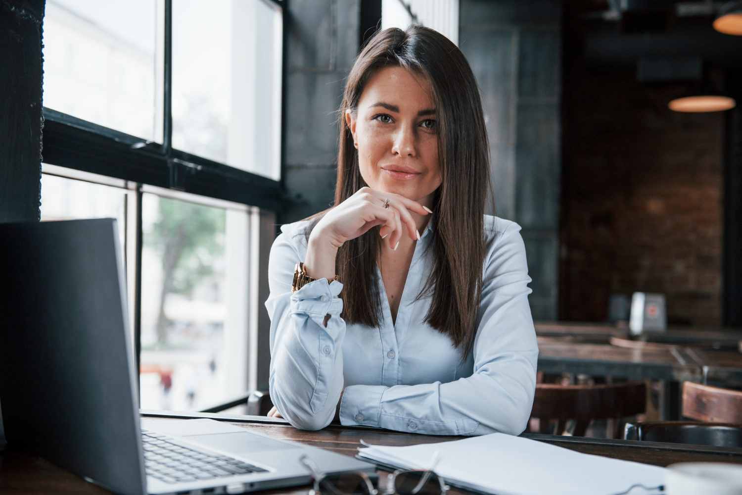 mulher com imagem profissional sorrindo pouco, com a mão no queixo. Ela está de cabelos soltos, com uma camisa social, sentada de frente para um notebook, com alguns documentos também em cima da mesa