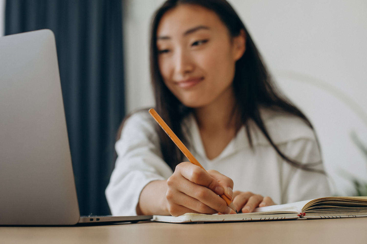 mulher escrevendo em um caderno a lápis enquanto olha para o notebook aberto sobre a mesa