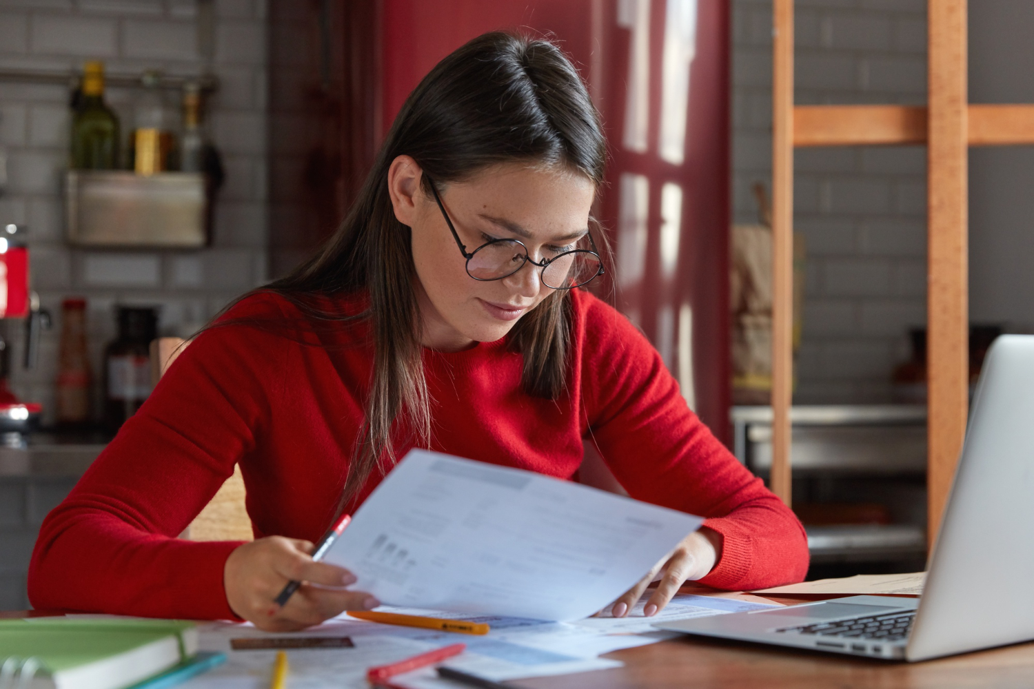 mulher olhando documentos sobre uma mesa de trabalho. A sua frente um notebook aberto
