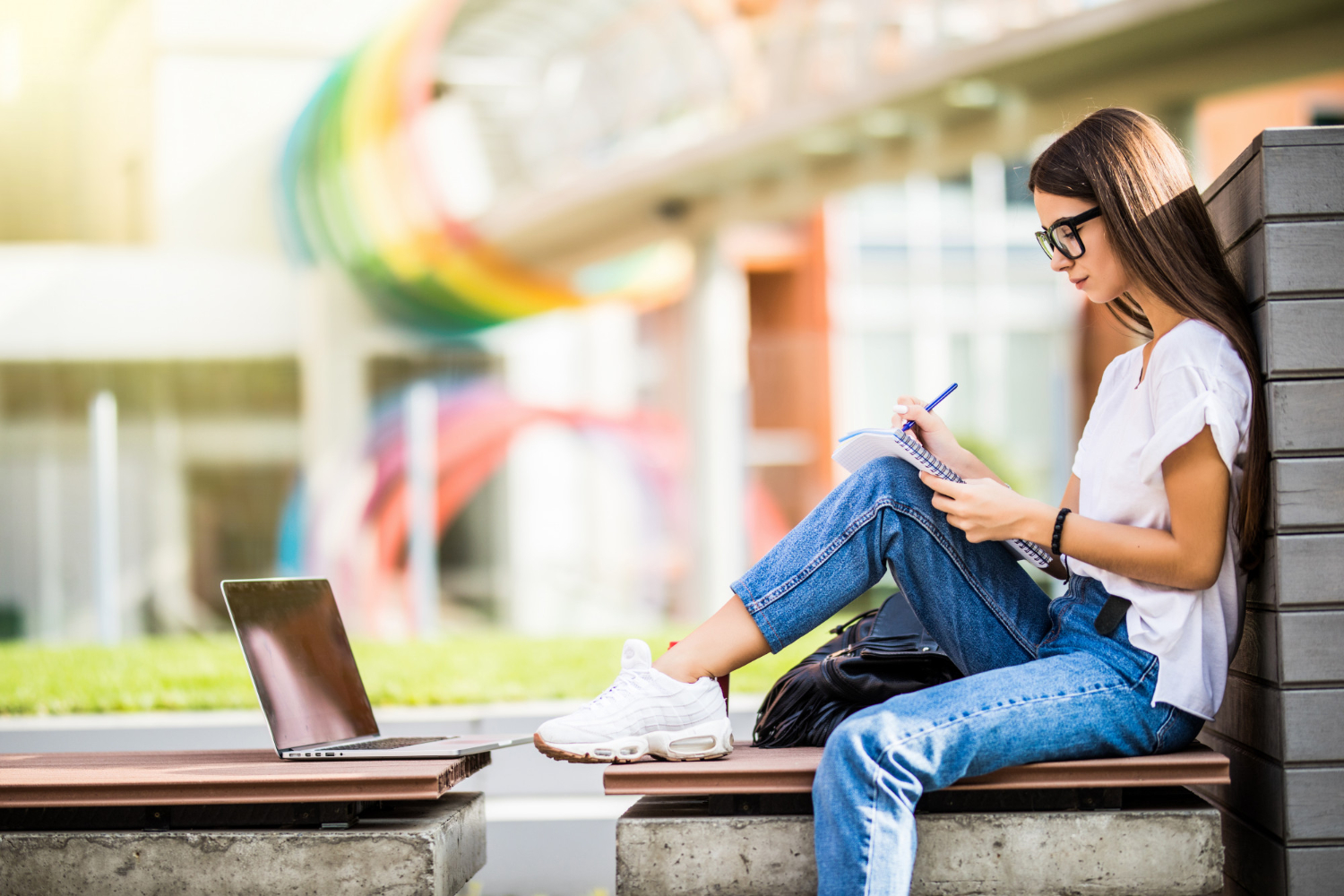 jovem adulta sentada na rua com cadernos e notebook de estudos. Ela está sentada com caderno sobre o joelho e seu notebook aberto em sua frente