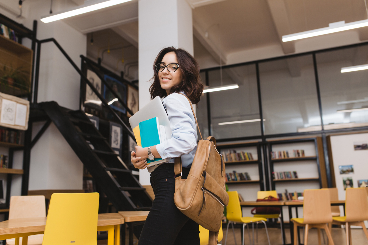jovem adulta em uma biblioteca de faculdade. Ela está em pé segurando notebook e livros, com a mochila sobre os ombros e está sorrindo para a câmera