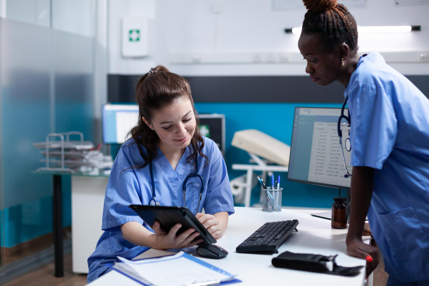 duas mulheres com parametrização de enfermagem em consultório. Uma delas está mostrando algo, em um tablet, para a outra.