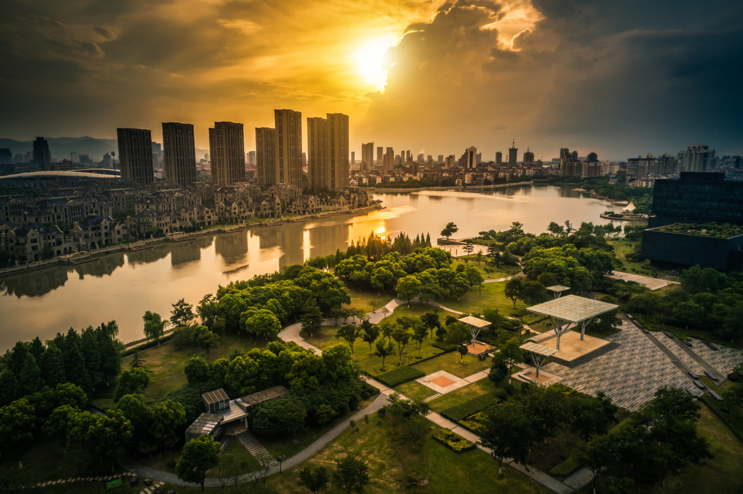 imagem aérea de uma cidade com gramado e árvores em primeiro plano, um rio ao meio e prédios ao fundo no pôr-do-sol. A imagem representa o que é urbanismo.
