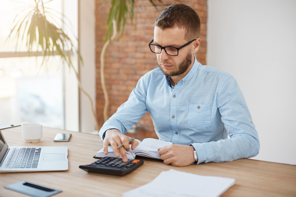 Homem em frente ao notebook com calculadora e papel.