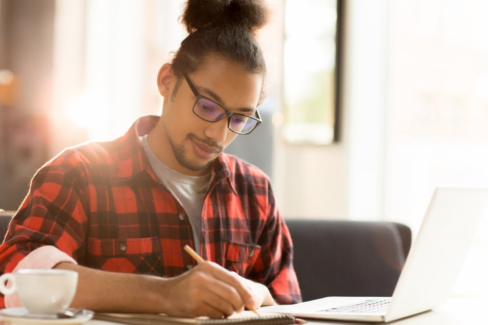 jovem adulto sentado estudando. Ele está fazendo anotações de estudos em um caderno que está ao lado do seu notebook