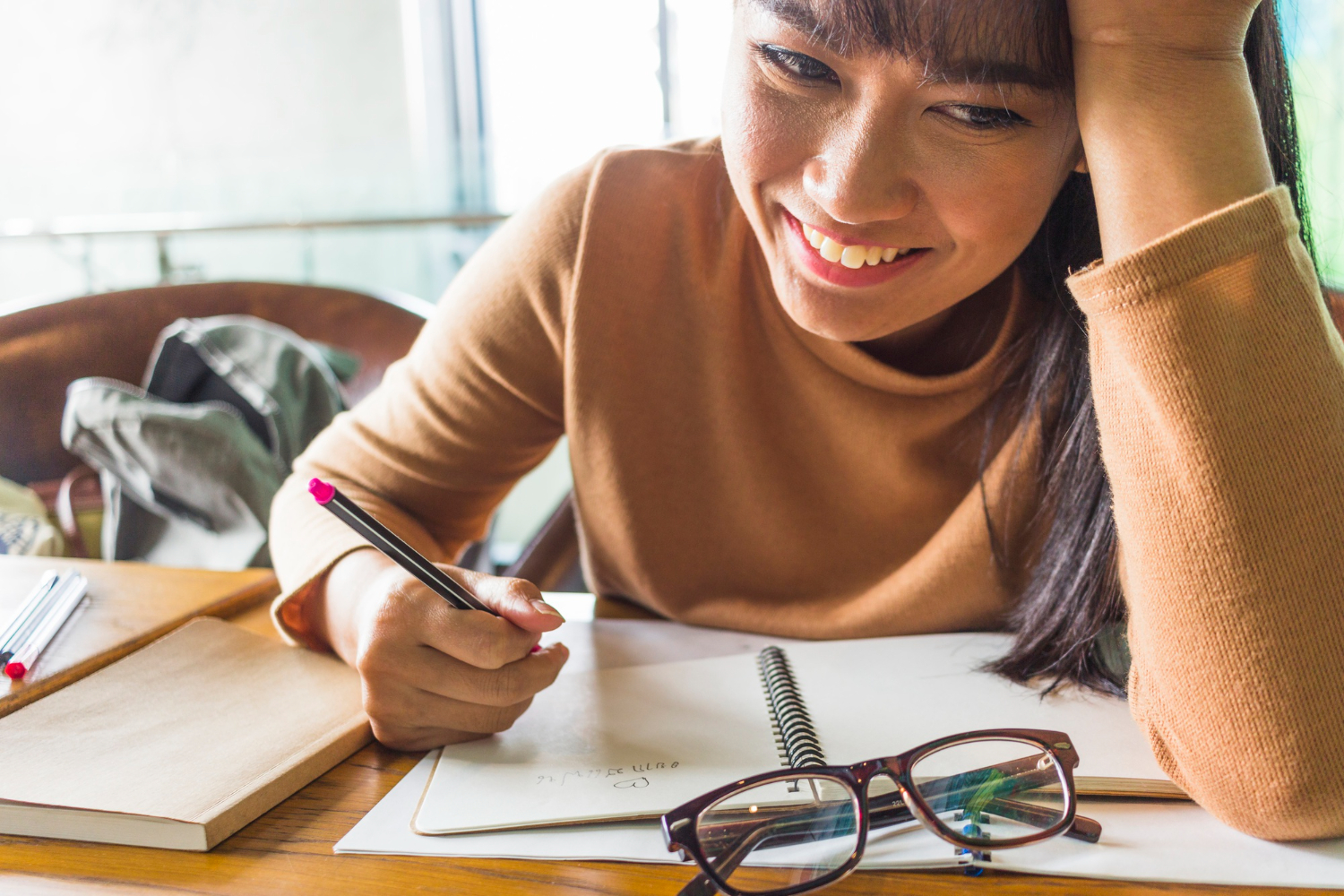 mulher sorrindo ao estudar. Ela está com cadernos abertos sobre a mesa, segurando uma caneta em uma das mãos e a cabeça apoiada na outra mão