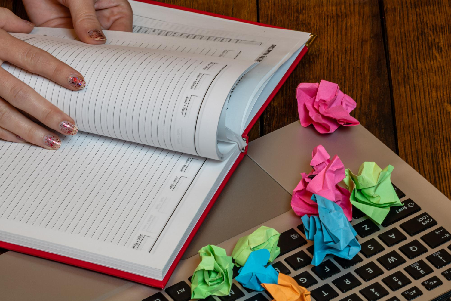 imagem vista de cima. Um caderno aberto com alguém folheando as páginas, está sobre um notebook. Em cima do notebook há também diversos postits amassados