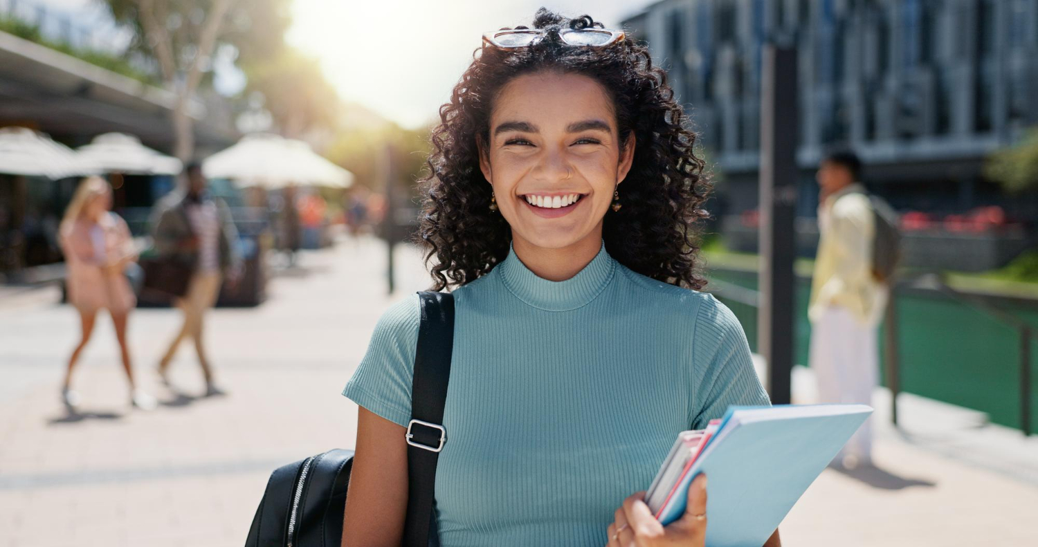 Jovem adulta segurando cadernos em uma das mãos, com uma mochila pendurada no ombro. Ela está sorrindo para a câmera e está em ambiente acadêmico, no lado de fora de uma faculdade
