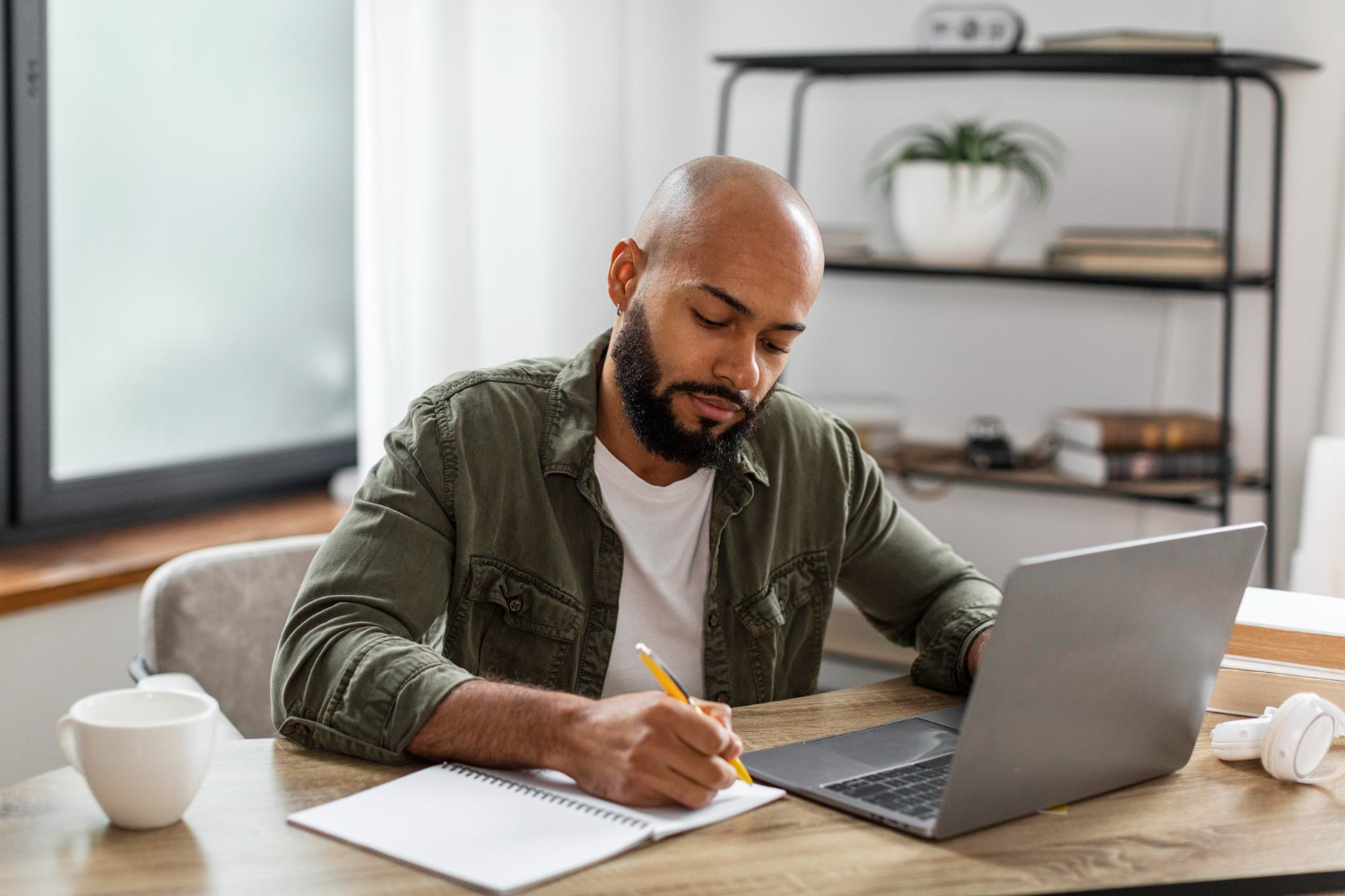 homem estudando em casa, na mesa. Está escrevendo em um caderno com uma das mãos e a outra está sobre o notebook aberto em sua frente