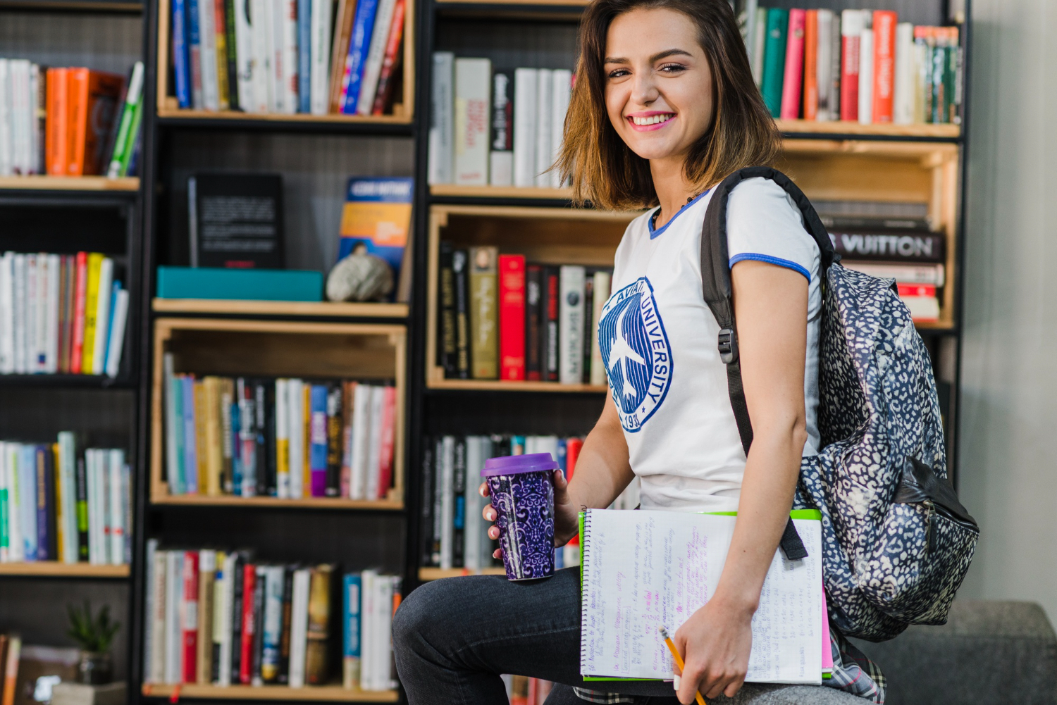 Menina morena segurando cadernos e caneca de cafe na biblioteca da faculdade sorridente